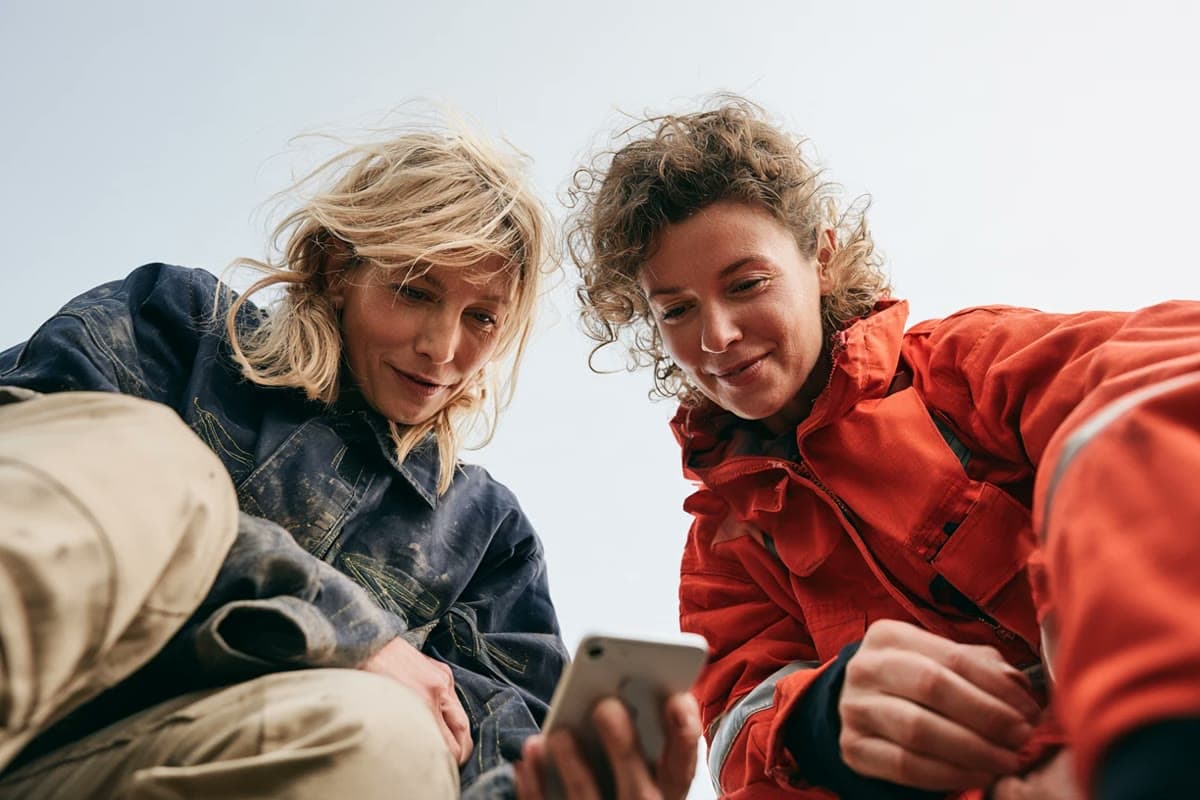 Dos trabajadoras de primera línea con chaquetas de trabajo, al aire libre, se inclinan juntas para mirar un móvil.